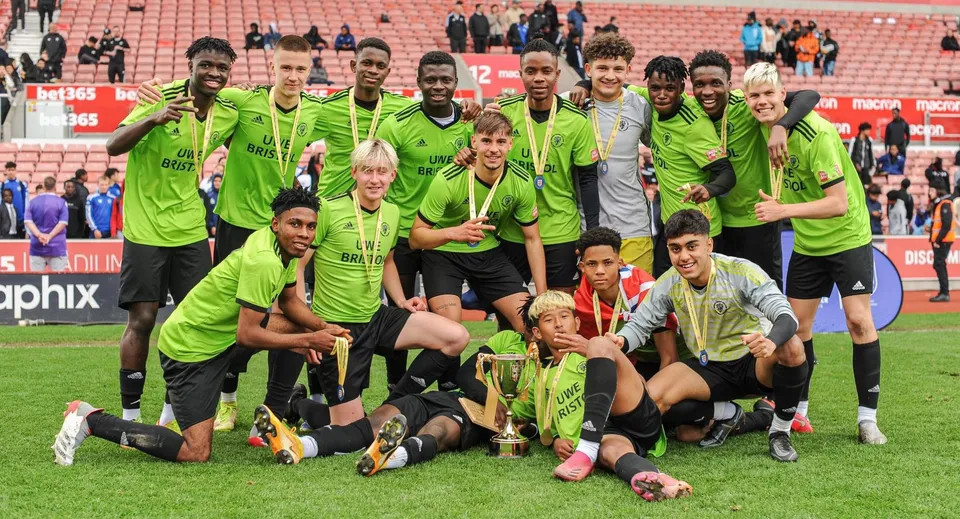 Studenti del Brooke House College festeggiano un gol sul campo da calcio con gli spettatori nello stadio sullo sfondo.