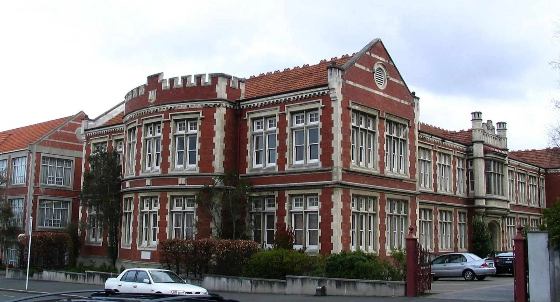 Il grande edificio in mattoni con architettura distintiva della Otago Girls High School è circondato da alberi e auto parcheggiate.