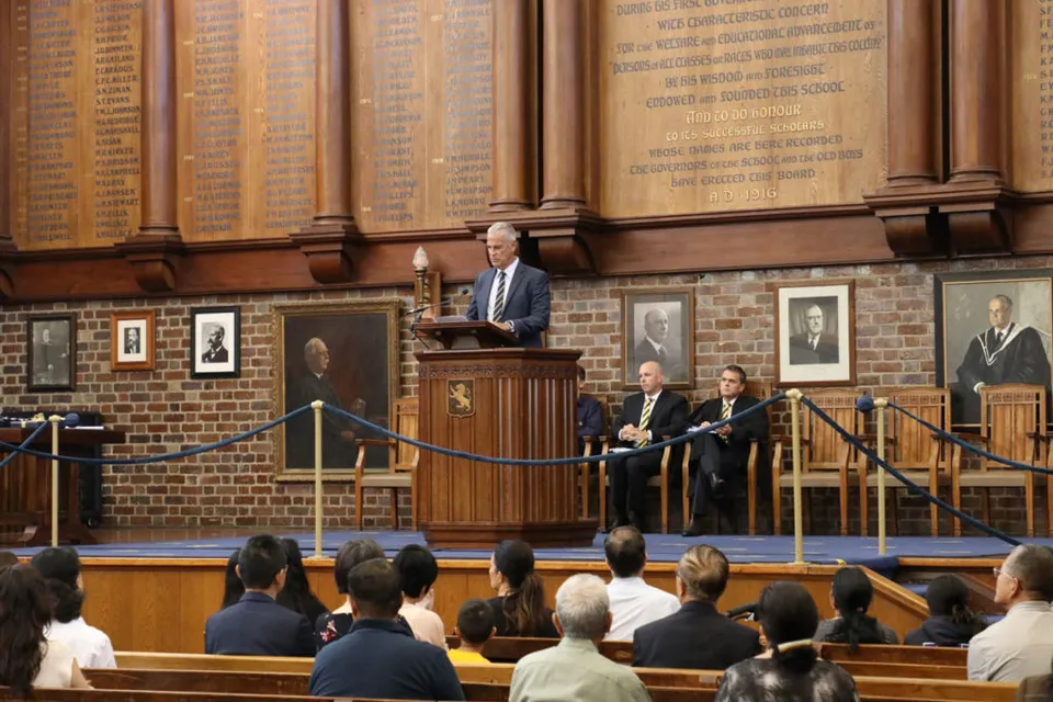 Un uomo si trova al leggio e parla davanti a un grande pubblico nella sfarzosa aula della Auckland Grammar School.