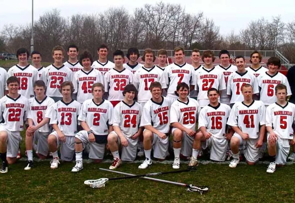 Un gruppo di studenti della Marblehead High School in divisa sportiva bianca e rossa si trova su un campo erboso con alberi sullo sfondo.