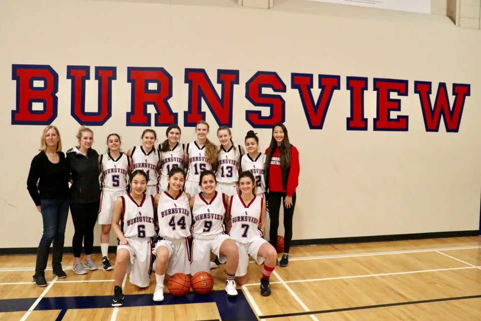 Un gruppo di giocatrici di basket posa davanti all'insegna della Burnsview Secondary School nella palestra.