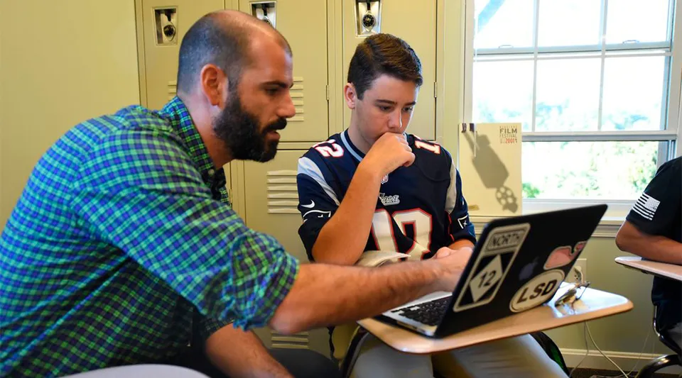 Un uomo con la barba e uno studente sono seduti insieme al laptop nel campus della Sandy Spring Friends School.