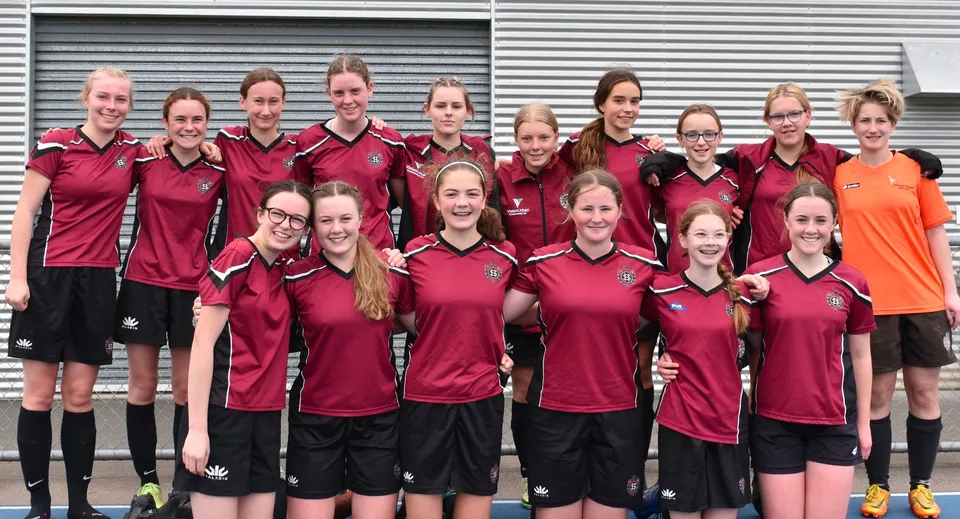 Un gruppo di studentesse del Sacred Heart College Napier in posa con uniformi sportive rosse e nere davanti a una tapparella metallica.