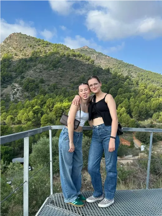 Zwei Mädchen posieren auf einer Plattform mit grünen Hügeln und einem Berg im Hintergrund in Spanien