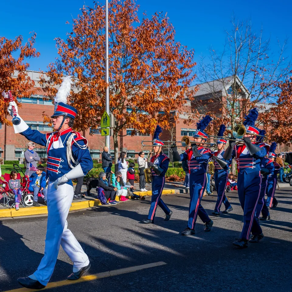 I membri della Marching Band della Kennedy Catholic High School si esibiscono in variopinte uniformi su una strada circondata da alberi autunnali.