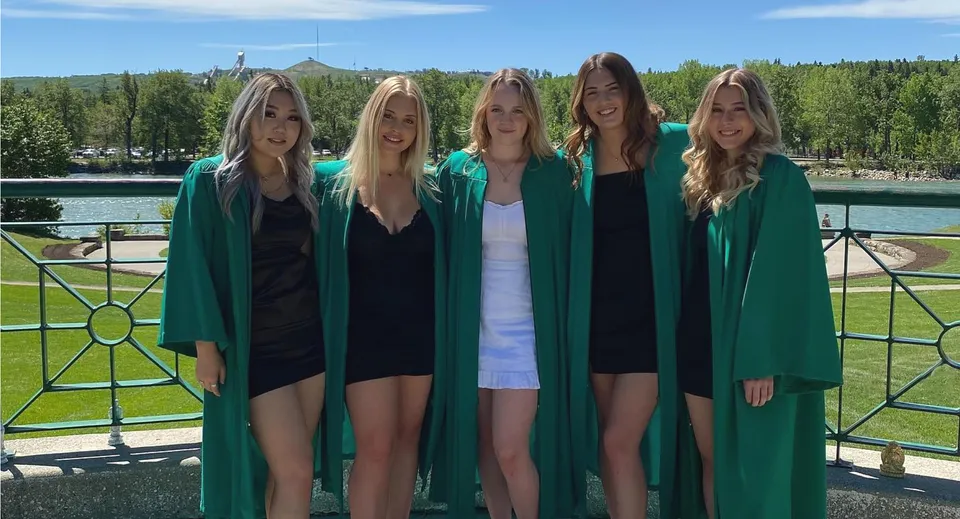 Un gruppo di cinque studentesse in toga verde da diploma si trova nel campus della Bowness High School davanti a un lago e alle montagne.