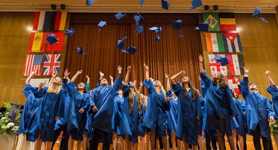 I neodiplomati della Glenlyon Norfolk School festeggiano in toga blu su un palco con bandiere internazionali.