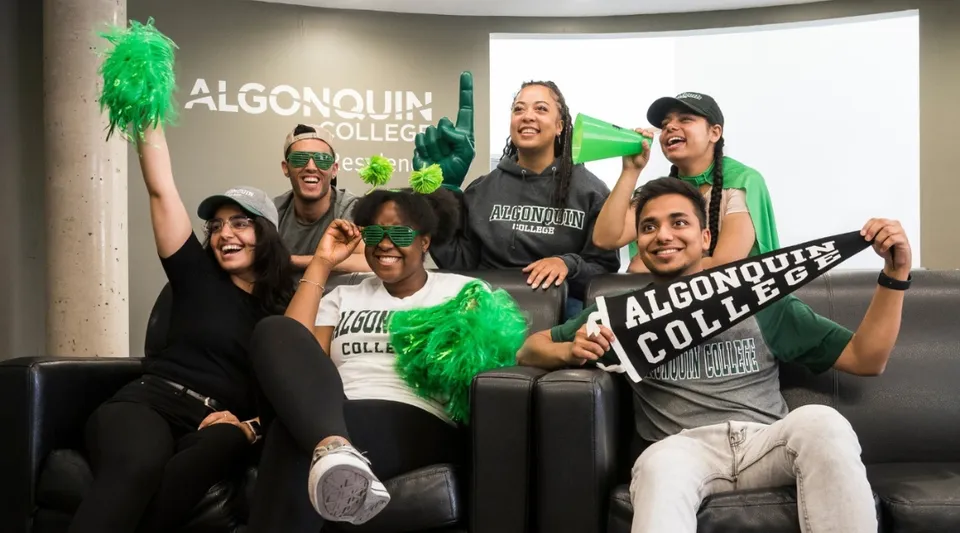 Un gruppo di studenti davanti al cartello dell'Algonquin College tiene dei pompon verdi e posa insieme.