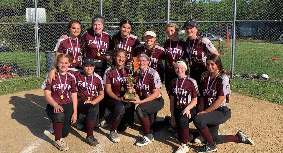 Un gruppo di giovani giocatrici di softball della Faith Christian Academy (PA) posa sul campo da baseball davanti a una recinzione a rete.
