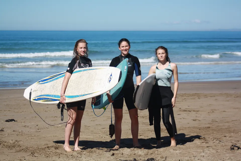 Tre studentesse della Gisborne Girls' High School in muta da sub con tavole da surf sulla spiaggia davanti all'oceano.