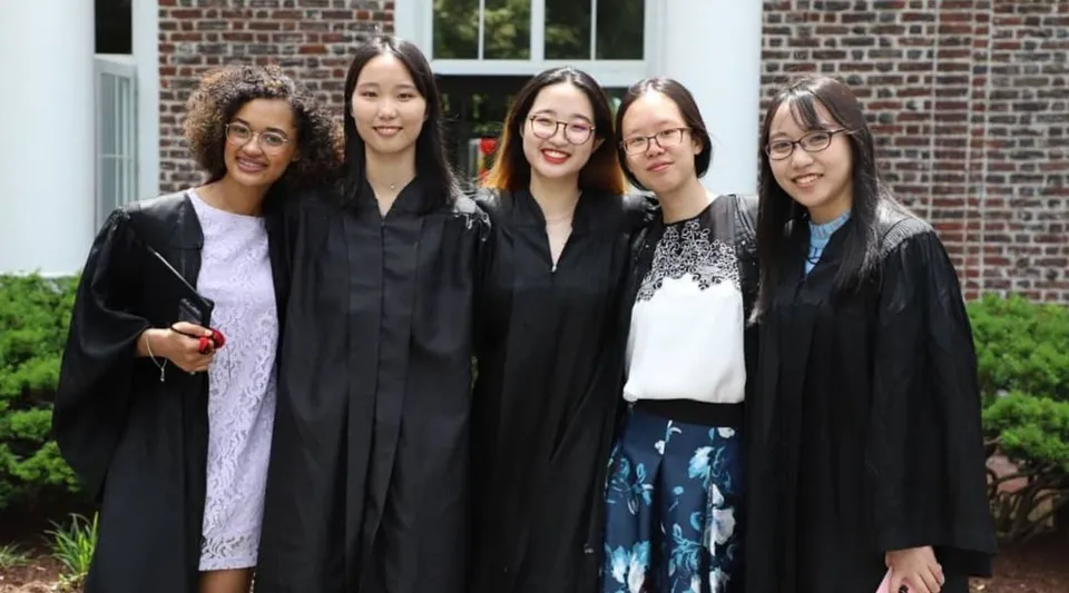 Cinque studentesse in toga da diploma sono in piedi davanti a un edificio in mattoni nel campus della Princeton International School of Mathematics & Science.