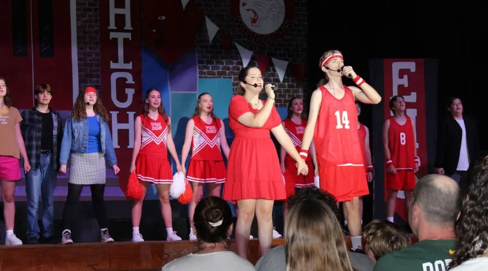 Un gruppo di studenti della Woolgoolga High School si esibisce in costumi da cheerleader rossi su un palco con diversi striscioni pubblicitari sullo sfondo.