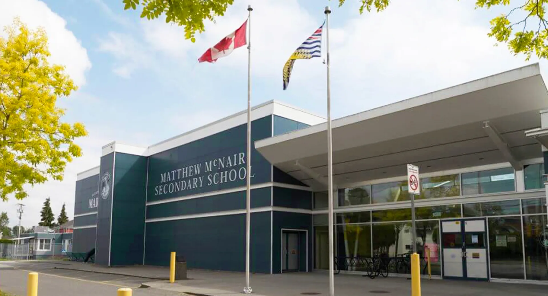 L'edificio scolastico moderno a più piani della Matthew McNair Secondary School è circondato da alberi e presenta diverse bandiere nell'area esterna.