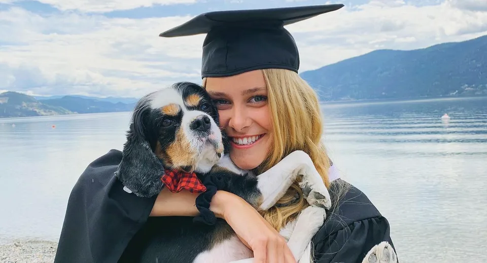 Una diplomata sorridente della George Elliot Secondary School tiene un cane sulla spiaggia con le montagne sullo sfondo.