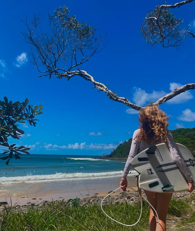 Chica de espaldas con una tabla de surf mirando hacia la playa entre los árboles