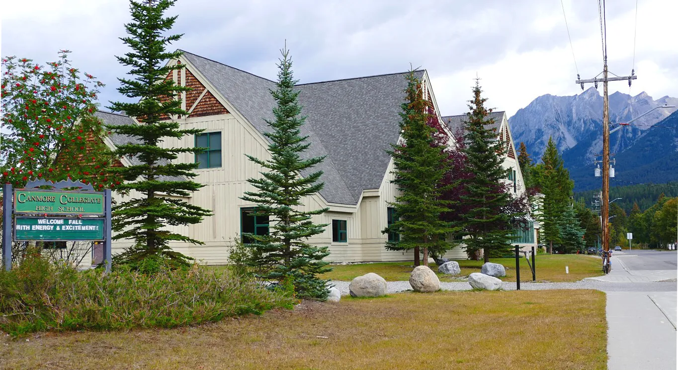 El edificio rústico de Canmore Collegiate High School está integrado en un paisaje boscoso con altos pinos y montañas al fondo.