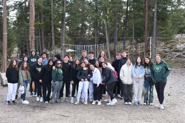 Un grupo de estudiantes de la Immaculata Regional School está junto en una zona arbolada con un puente colgante al fondo.