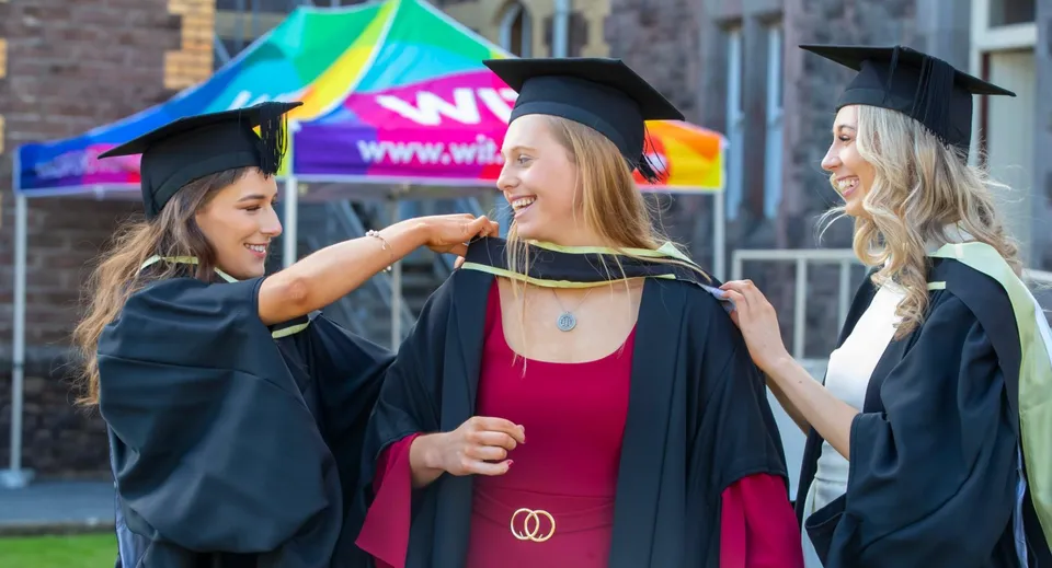 Tre studentesse della Kilkenny City Vocational School festeggiano il loro diploma davanti a uno sfondo colorato con un cartello WIN.