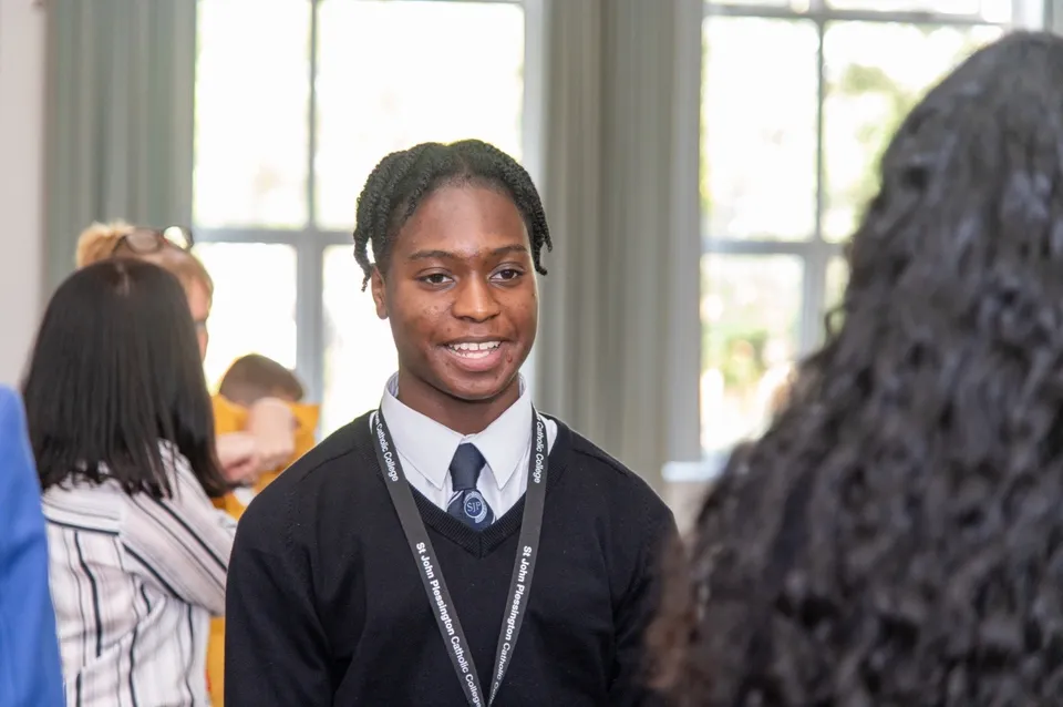 Uno studente in divisa scolastica sorride in un'aula del St. John Plessington Catholic College con altri studenti sullo sfondo.