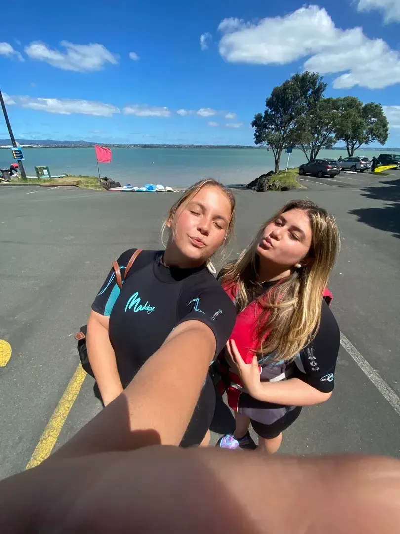 Zwei Mädchen in Neoprenanzügen machen ein Selfie an einer Küstenstraße mit Bucht und Bäumen im Hintergrund