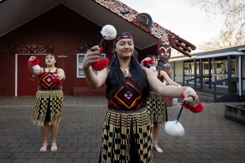Due studentesse della Hamilton Girl's High School indossano abiti tradizionali Maori davanti a un edificio caratteristico della scuola.
