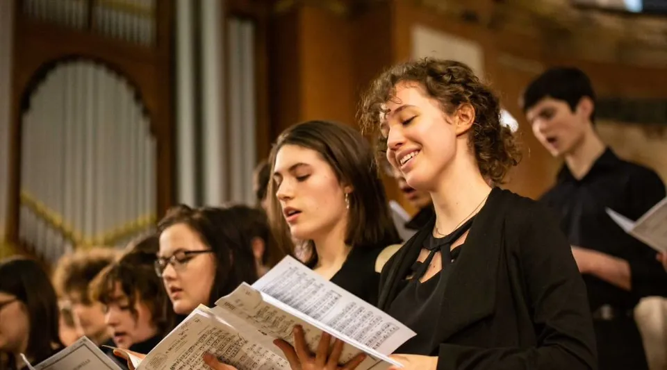Un gruppo di studenti della St. Mary’s Music School canta insieme su un palco con uno sfondo in legno.