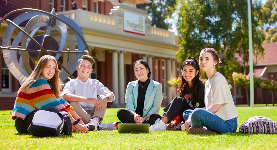 Un gruppo di studenti è seduto sul prato davanti a un edificio in mattoni con una grande ruota nell'area del Thebarton Senior College.
