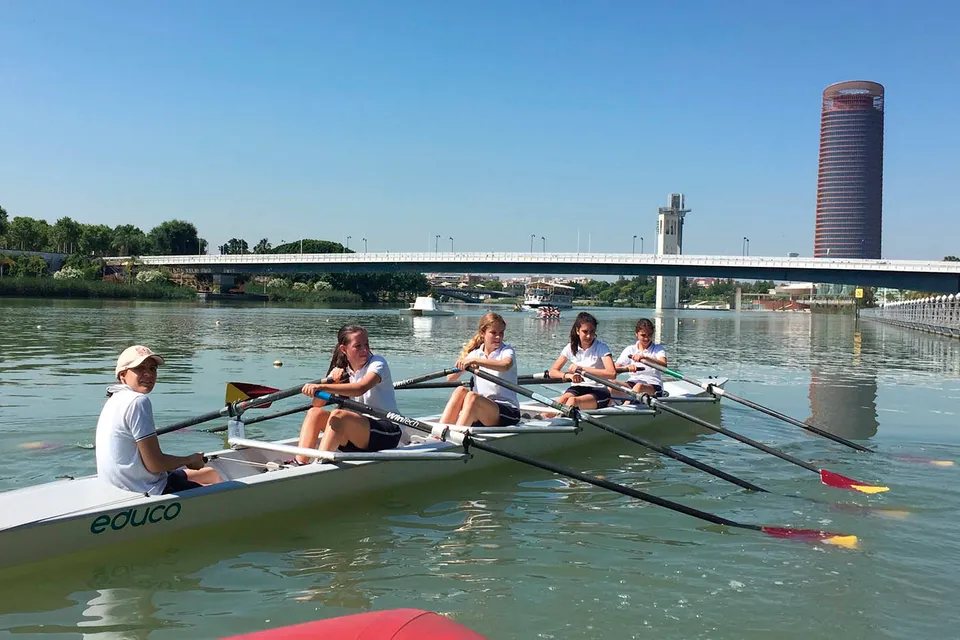 Studenti della Yago School fanno canottaggio su un fiume con un ponte e un'alta torre sullo sfondo.