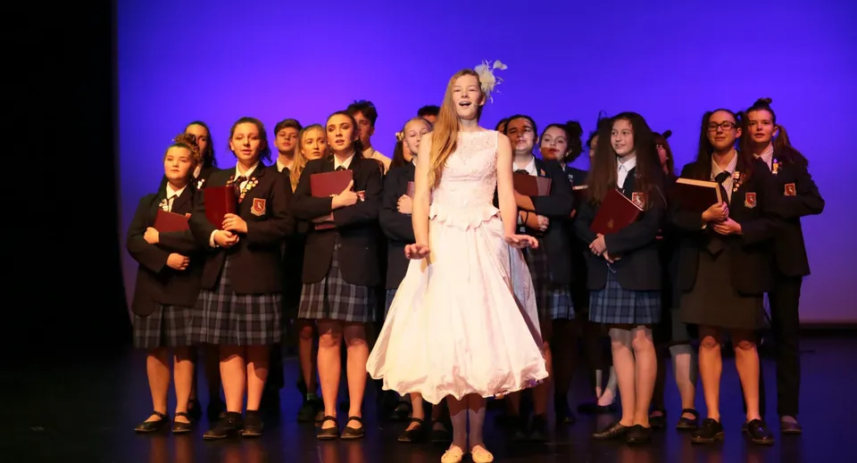 Una donna in abito bianco si trova sul palco davanti a un gruppo di studentesse nel campus della St. Peter's School Cambridge.