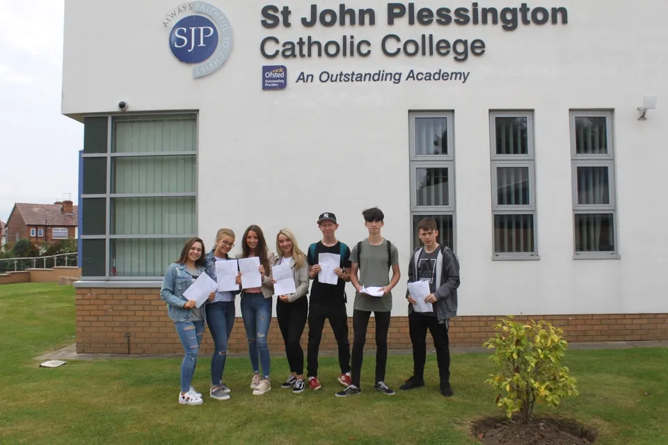 Un gruppo di studenti e studentesse si trova con dei certificati davanti all'edificio del St. John Plessington Catholic College.