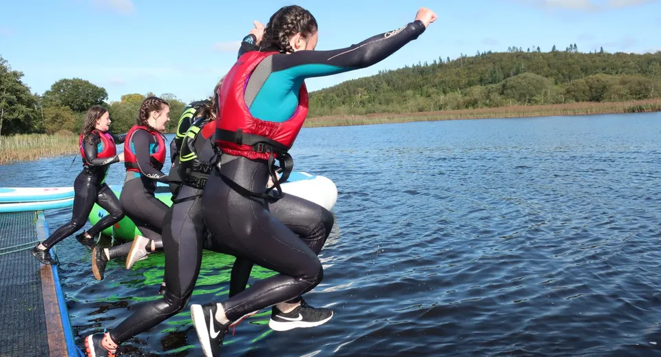 Studentesse del Castleblayney College con mute e giubbotti salvagente saltano in un lago da un pontile.