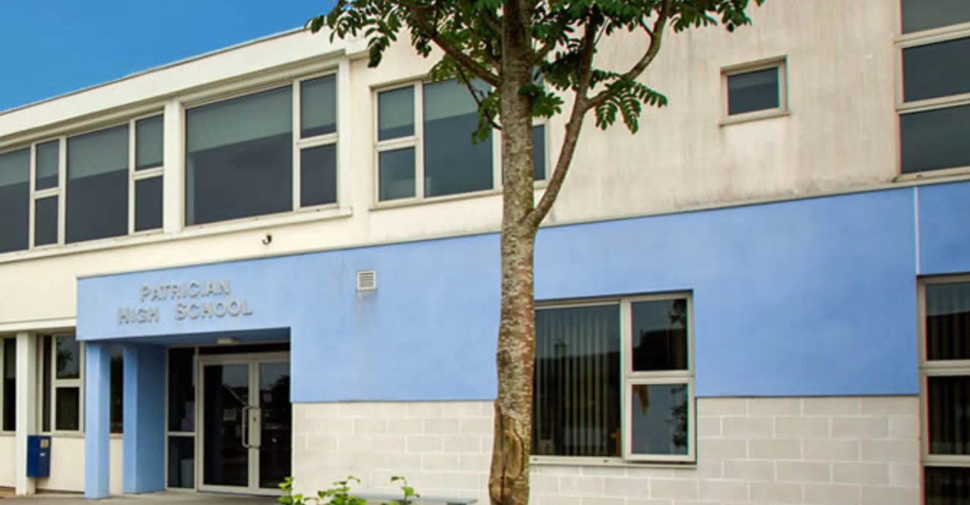 L'edificio moderno a due piani della Patrician High School con grandi finestre e un albero in primo piano sotto un cielo limpido.