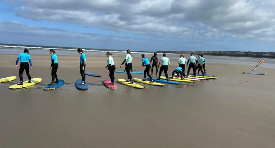 Studenti del Davitt College partecipano a un corso di surf sulla spiaggia sotto un cielo nuvoloso.