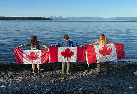 Tres jóvenes de espaldas sujetando banderas de Canadá a la orilla de un lago