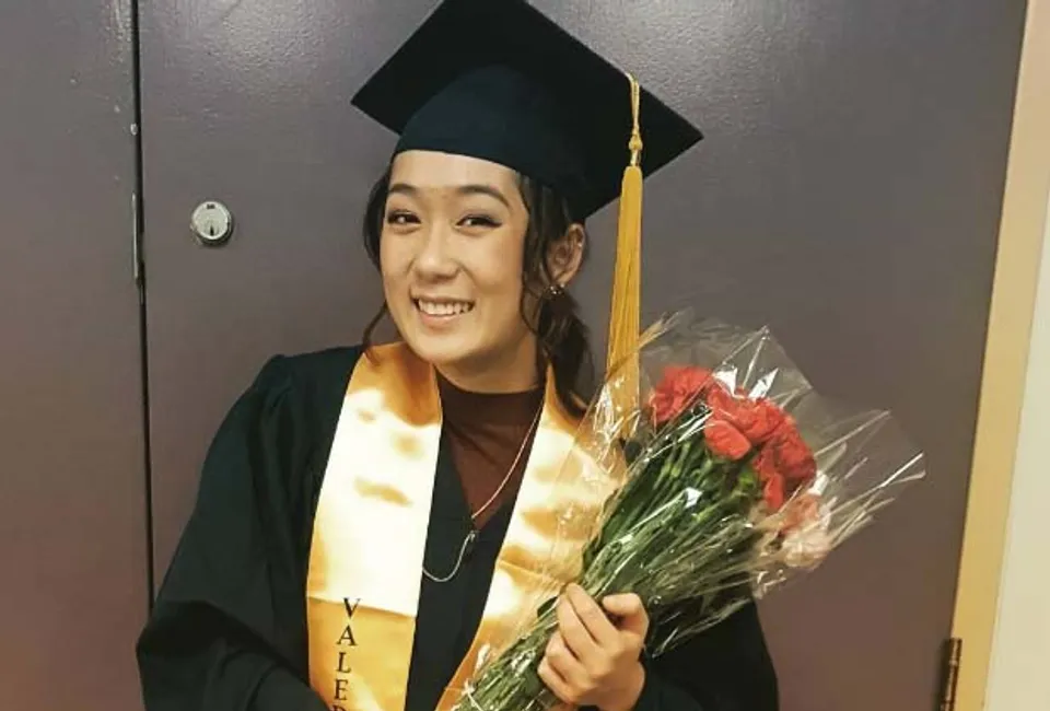 Una diplomata della Central Technical School in toga tiene un mazzo di rose rosse davanti a una porta e sorride.