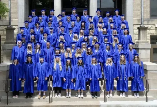 Un gran grupo de graduados de Cotter Schools está con togas y birretes azules en las escaleras de un edificio con fachada de piedra.