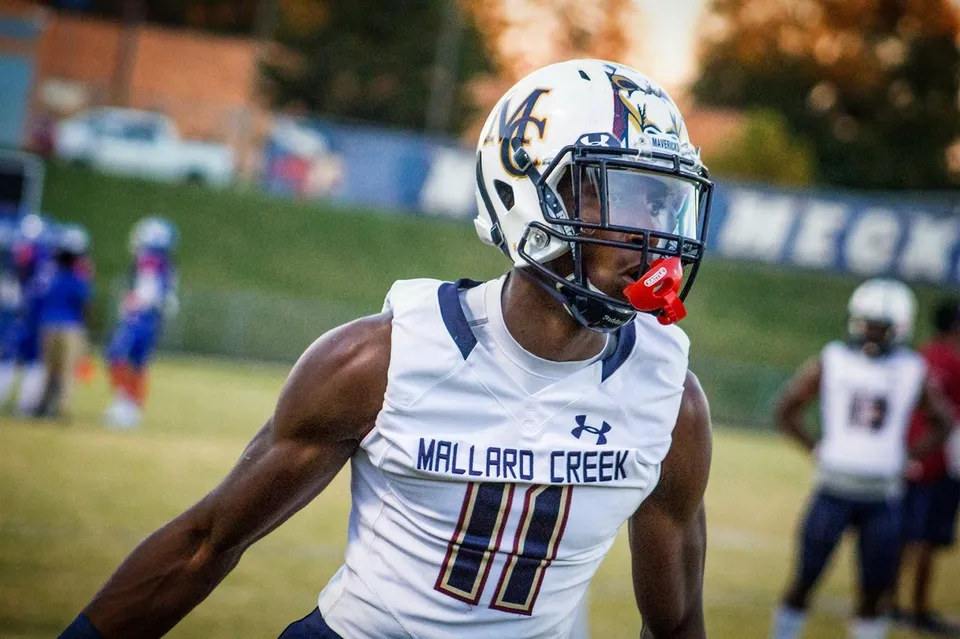 Un giocatore di football in uniforme bianca della Mallard Creek High School si trova sul campo con altri giocatori e spettatori sullo sfondo.