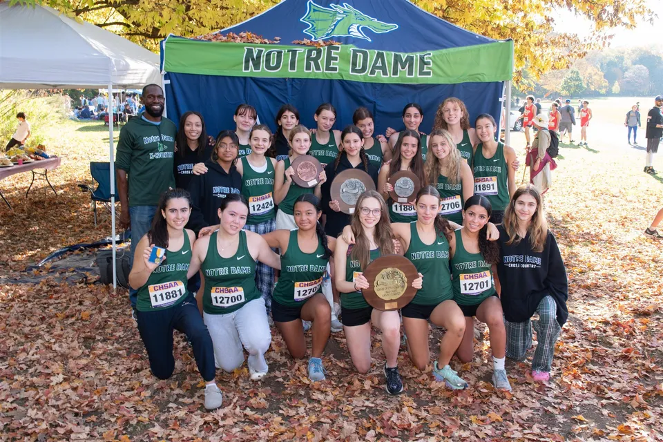 Un gruppo di atlete con maglie verdi e bianche posa davanti a uno striscione della Notre Dame High School in un bosco autunnale.