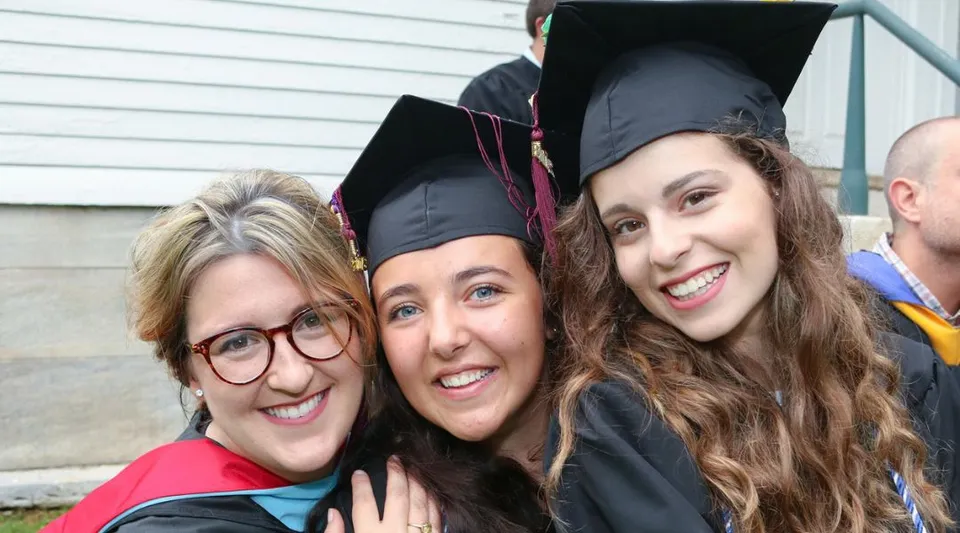 Tre giovani donne in abito da diploma della Darrow School si trovano davanti a un edificio in legno.