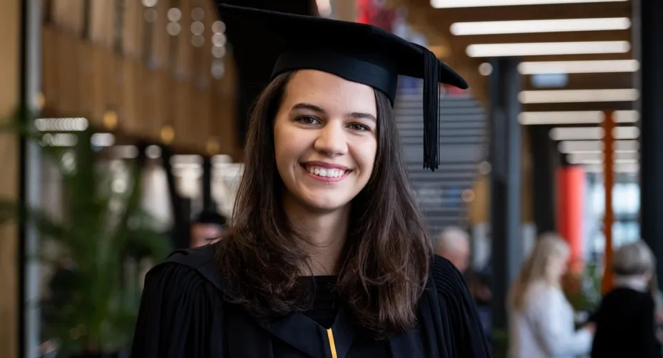 Una neodiplomata sorridente in toga nera sta davanti a una struttura in legno decorata nel campus del Tauranga Girl´s College.