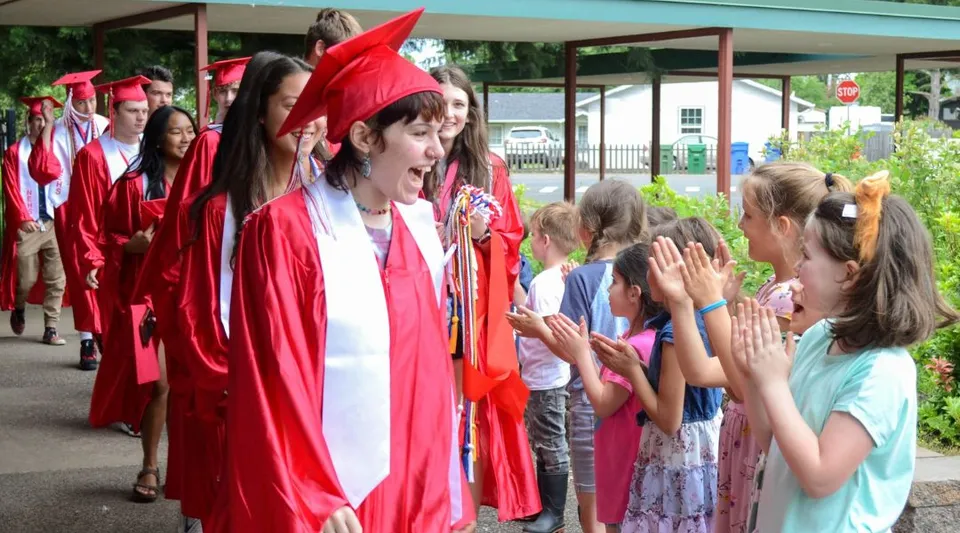 Studenti del Eugene School District in toga rossa camminano davanti a bambini che applaudono.