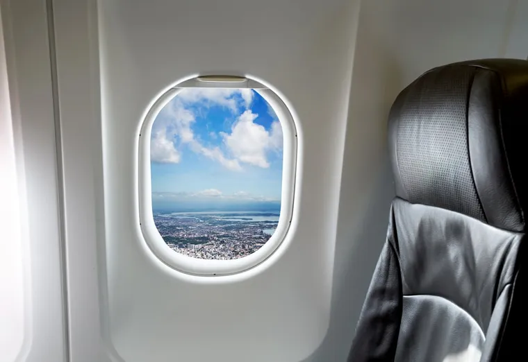 Blick durch ein Flugzeugfenster auf Wolken und eine Stadt, daneben ein Ledersitz