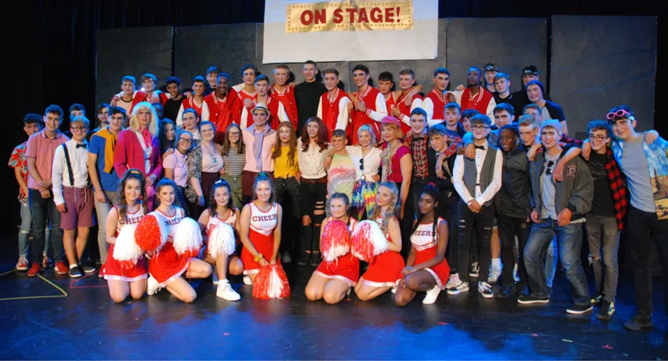 Un gruppo di studentesse e studenti della St. Joseph’s Secondary School si trova su un palco in costumi colorati con un cartello 'On Stage!' sullo sfondo.