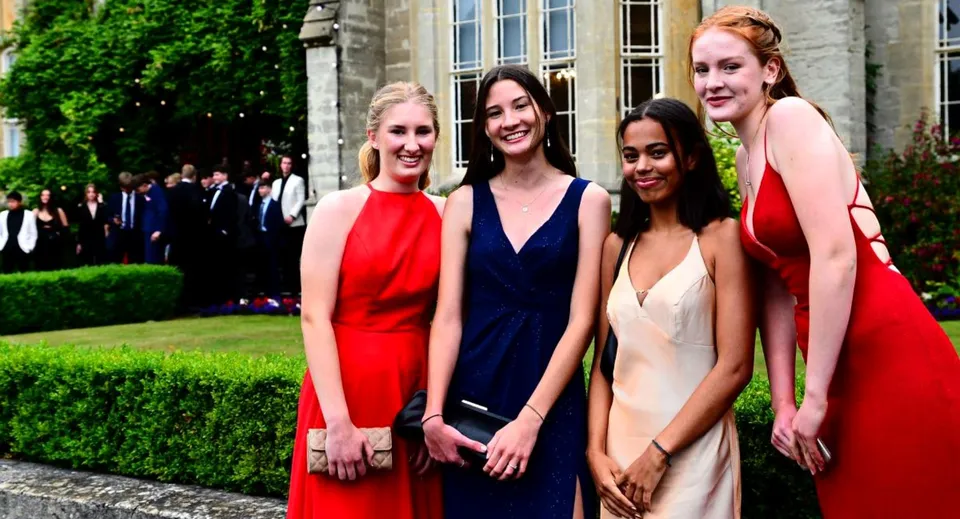 Quattro studentesse del Queen’s College in abito formale si trovano insieme in un giardino con un edificio sullo sfondo.