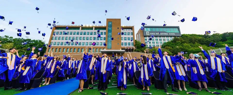 Eine große Gruppe von Absolventen der Korea International School steht vor einem Universitätsgebäude und wirft ihre blaue Abschlussmütze in die Luft.