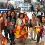 Grupo de personas en un aeropuerto posando sonrientes mientras sostienen varias banderas de España