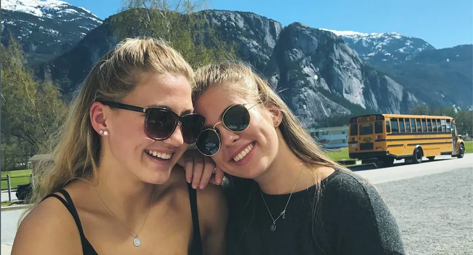 Due studentesse con i capelli biondi posano davanti a un paesaggio montano nel campus della Howe Sound Secondary School.