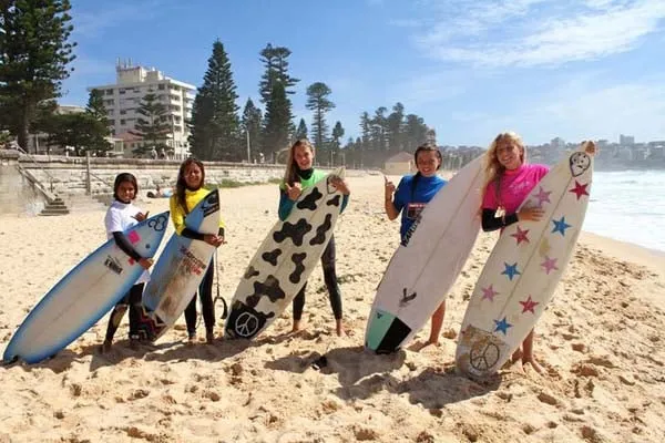Alumnas del Mackellar Girls Campus están de pie con tablas de surf de colores en la playa frente a edificios y árboles.