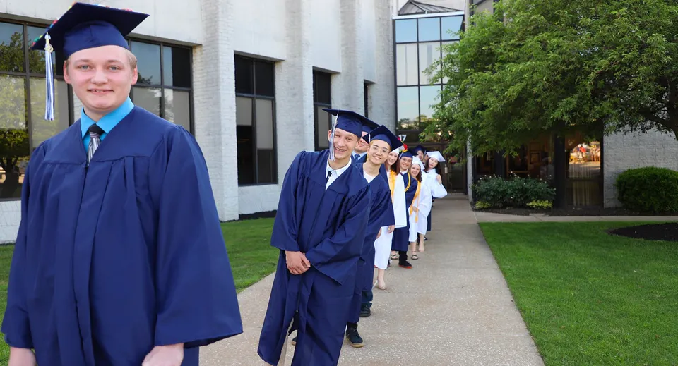 Un gruppo di diplomati in toga e tocco blu si trova nel campus della Erie First Christian Academy.
