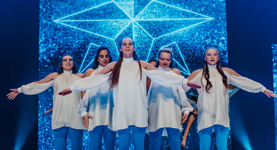 Un gruppo di studentesse della Faith Christian Academy (FL) esegue un'esibizione di danza sul palco davanti a uno sfondo geometrico illuminato.
